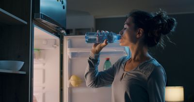 A woman drinking water from a bottle in front of an open refrigerator.