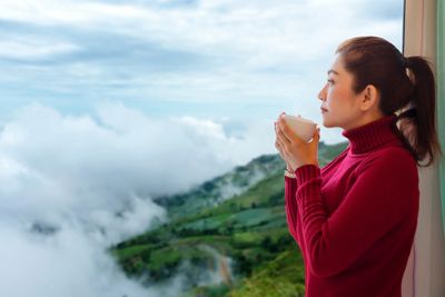 A woman in a red sweater standing next to the window looking over hills and holding a glass of water to relieve migrainePerson holding a glass of water in a tranquil nature setting