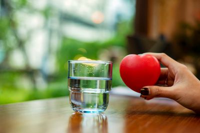 A hand holding a red heart next to a glass of water on a wooden table