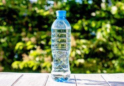 Image with vivid colours - a bottle of spring water (blue hues) on a white table, with lush greenery faded out in the background.