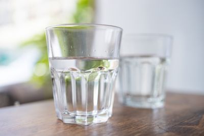 A zoom-in on a tumbler glass about 2/3 filled with water, with another glass in the background, on a brown wooden table.