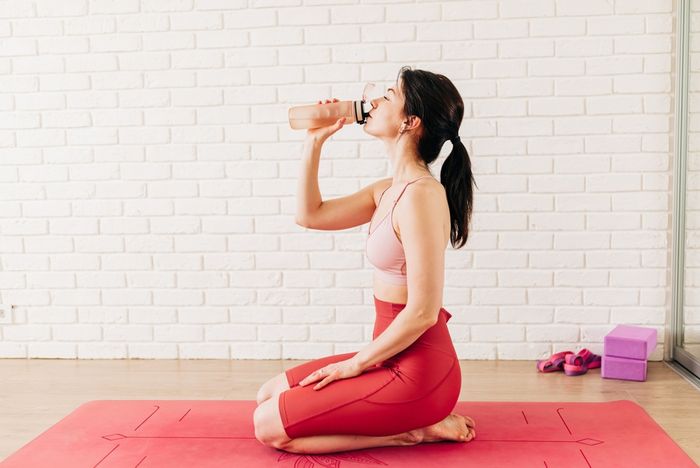 Woman sitting on a yoga mat drinking water from a glass bottle