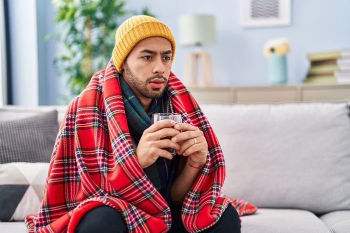 A sick man sitting on a couch wrapped in a blanket with a glass of water.