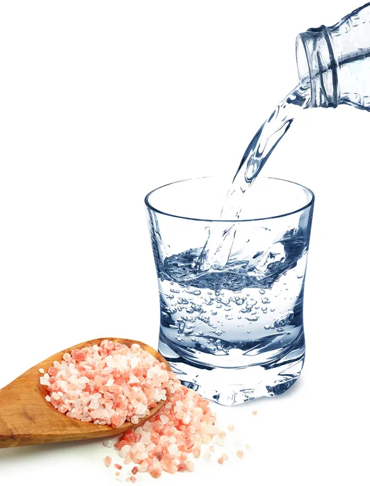 Water being poured from a bottle into a glass next to a wooden spoon holding Himalayan salt