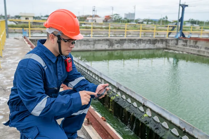 Man in overalls and hardhat looking at smartphone while crouching next to body of water