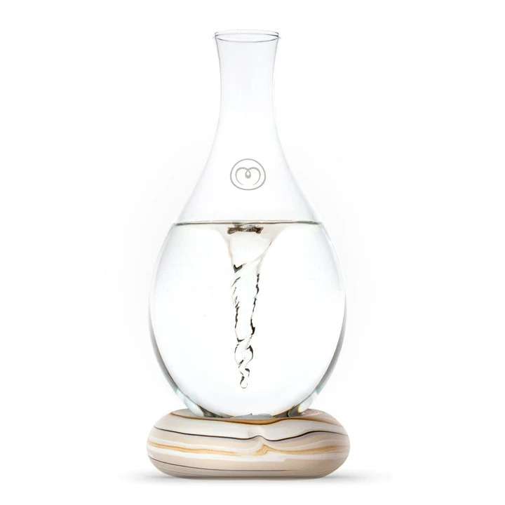 A stock image of a glass water pitcher with water swirling inside on a base in an earthy brown tone